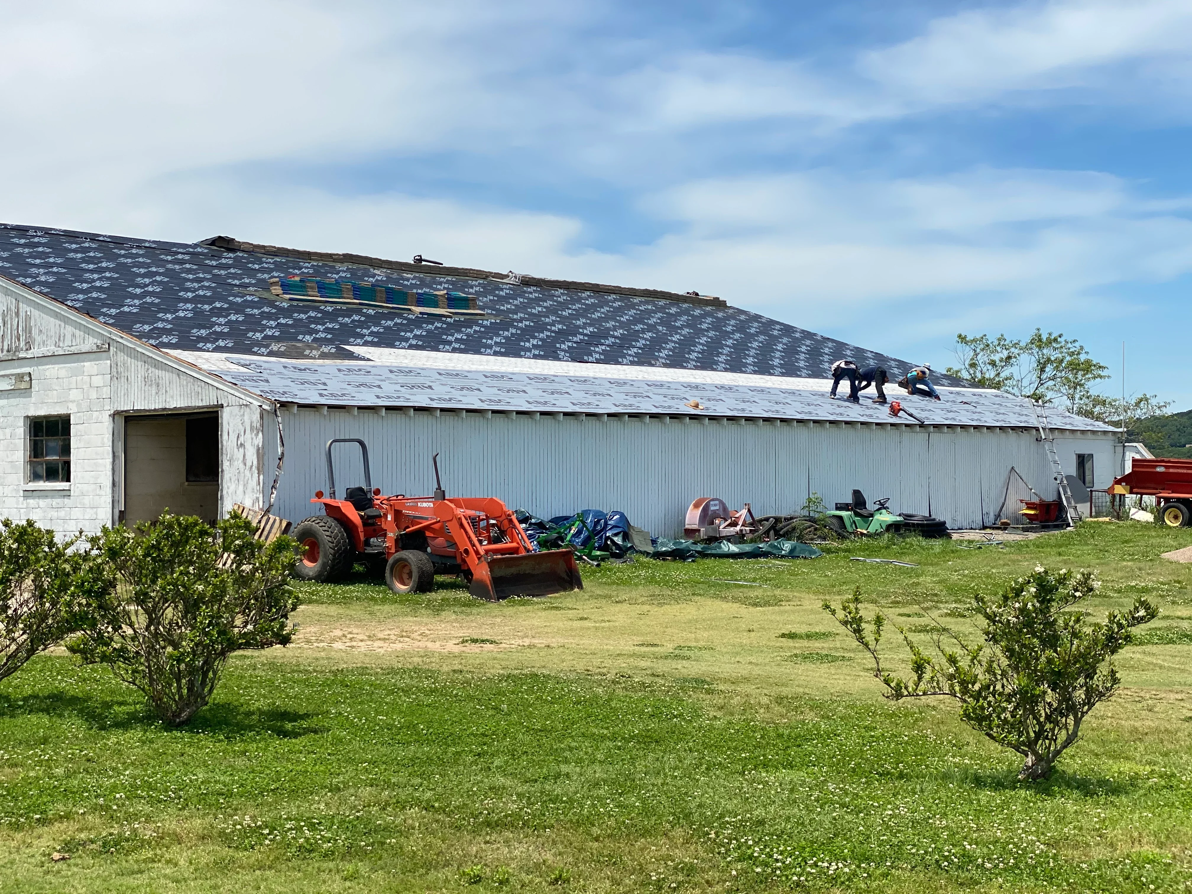 Metal agricultural building with new asphalt shingle roof installation in progress, workers on roof, white corrugated siding