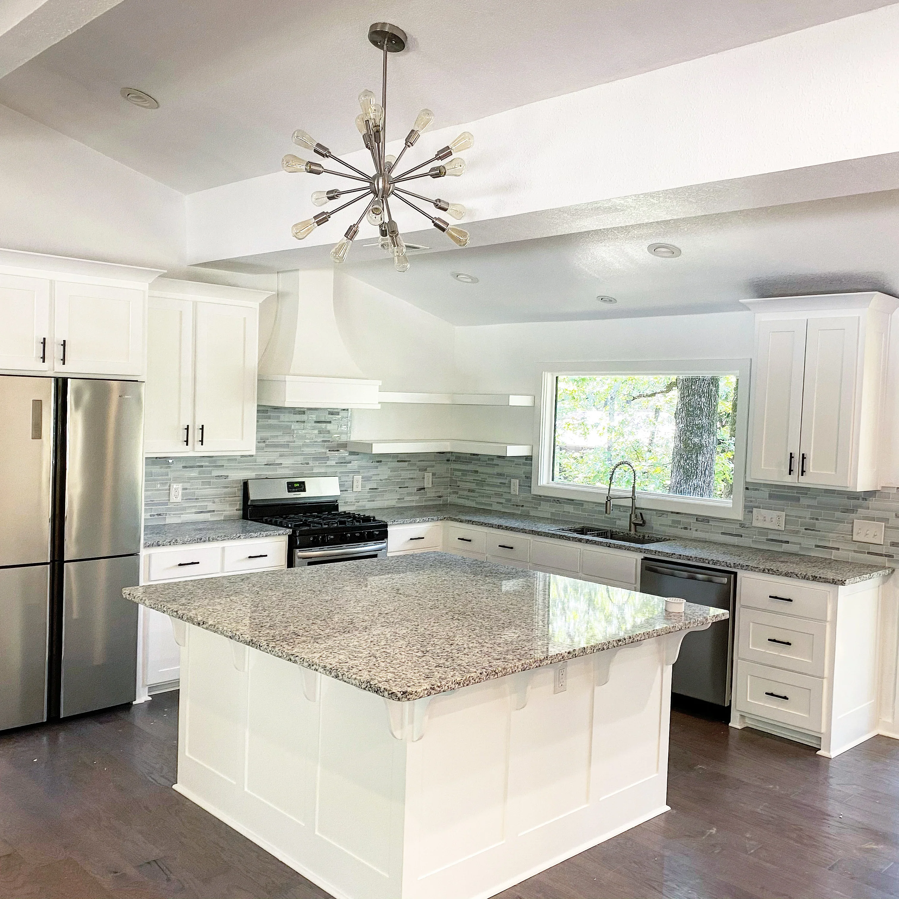 White kitchen with granite island, glass tile backsplash, stainless steel appliances, and modern lighting